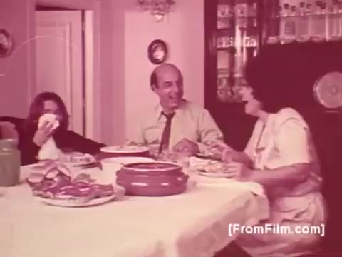 The still features a dining scene from a 1970s Knorr Caldo De Galinha soup commercial in Brazil. The setting is a homely kitchen or dining area, with a large table laid out with bowls of food, likely including the soup highlighted in the ad. Three characters are present: a woman serving food, a man smiling at her, and a child playfully covering their face. The overall atmosphere is warm and familial, emphasizing the joy of shared meals. The color tone suggests a vintage feel characteristic of that era.