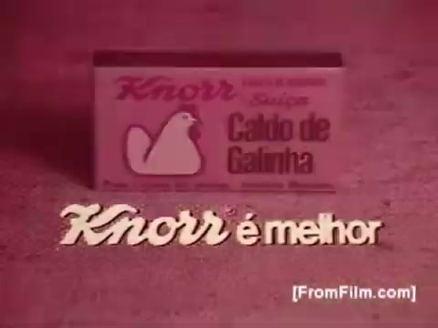 The still features a vintage 16mm print of a Knorr Caldo De Galinha soup commercial from the 1970s in Brazil. The focal point is a package of Knorr's chicken broth, prominently displayed with a simple design showcasing a chicken icon. The text 'Knorr é melhor' translates to 'Knorr is better,' emphasizing the product's quality. The overall color tone is warm and nostalgic, characteristic of the era's advertisements.