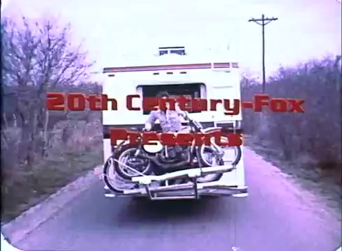 The still from the trailer of 'Race with the Devil' features a scene showing the rear of a motorhome driving down a rural road. The motorhome is equipped with bicycles strapped on the back. Prominently displayed in red is the logo '20th Century-Fox Presents,' indicating the production studio. The visual hints at adventure and the outdoor setting typical of the film’s themes.