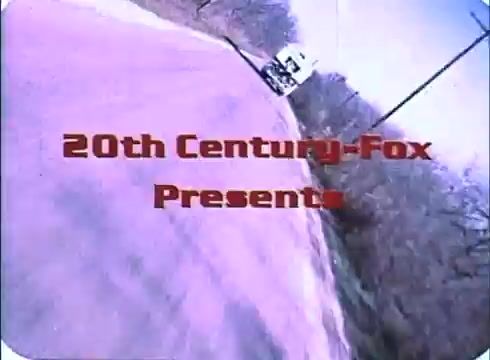 The still features a 16mm print from the trailer of 'Race with the Devil,' showcasing the title '20th Century-Fox Presents' prominently. It depicts a winding road, suggesting a sense of motion and adventure typical of the film's themes. The color palette has a slightly faded, vintage quality, indicative of the film's era. The truck in the background hints at the journey and potential conflicts central to the plot.