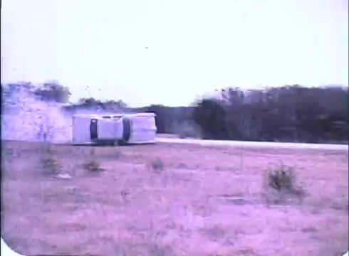 The still captures a scene from the trailer of 'Race with the Devil.' It shows a vehicle, likely a camper, overturned on the roadside, indicating a tense moment or crash. The background features sparse vegetation and a dusty landscape, emphasizing the isolation of the setting. The image has a vintage, slightly faded quality typical of 16mm prints.