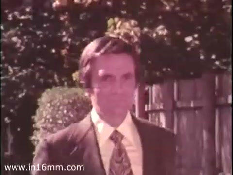 The still from the 16mm film 'Sole Leather Council Bicentennial PSA - 1975' features a man dressed in a suit walking towards the camera, with a neatly manicured hedge and a wooden fence in the background. The poster's message humorously advocates for using one's own feet ('shoe leather') over driving to save gas, emphasizing a practical and eco-friendly lifestyle choice. The overall tone is light-hearted and promotional.