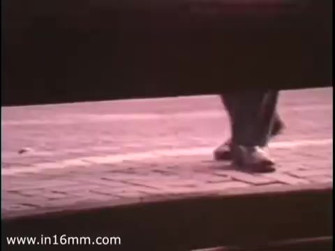 The still from 'Sole Leather Council Bicentennial PSA - 1975' features a close-up view of a person's legs as they walk on a brick surface. The scene emphasizes the message of using one's own foot power—'shoe leather'—promoting walking as an alternative to driving, thereby encouraging gas conservation. The aesthetic reflects the era's style, typical of 16mm film quality.