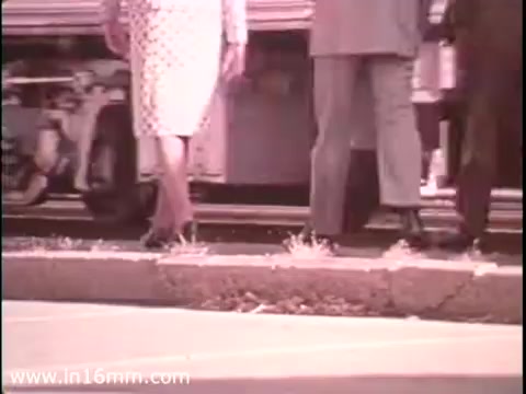 The still from the 'Sole Leather Council Bicentennial PSA - 1975' features three pairs of legs, highlighting a woman in a polka-dotted dress and two men in suits, as they walk along a sidewalk. The background suggests a bus or vehicle, emphasizing the theme of reducing gasoline consumption by promoting walking. The overall tone is lighthearted and promotes the message of using 'shoe leather' for transportation.