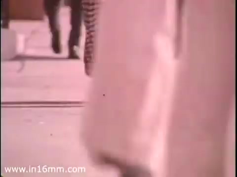 The still from 'Sole Leather Council Bicentennial PSA - 1975' captures a bustling street scene, focusing on people's legs and shoes as they walk on a sidewalk. The message conveyed is a playful promotion of walking as an eco-friendly alternative to driving, with the tagline 'Save gas! Use shoe leather instead!' The visual style reflects the era, likely featuring muted colors typical of 16mm film from that time.