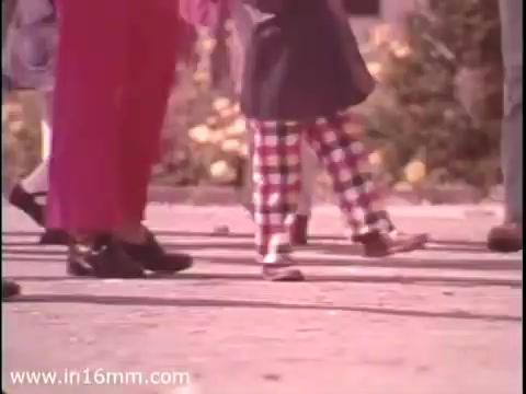 The still from 'Sole Leather Council Bicentennial PSA - 1975' features a close-up of feet walking on a pavement. Various shoes are visible, including smart dress shoes and sneakers, alongside a child in checkered pants. The image emphasizes the theme of promoting walking over driving, aligning with the PSA's message to 'save gas' by using 'shoe leather' instead. The setting includes greenery and flowers, adding a lively backdrop to the scene.