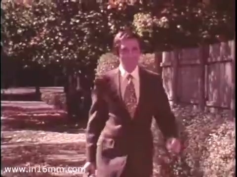 The still from 'Sole Leather Council Bicentennial PSA - 1975' features a man walking along a tree-lined sidewalk. He is dressed in a suit with a patterned tie and holds a newspaper. The vibrant colors of the foliage suggest a sunny day, highlighting the message to promote walking and saving gas by using 'shoe leather.' The setting conveys a leisurely, community-oriented atmosphere typical of public service announcements from that era.