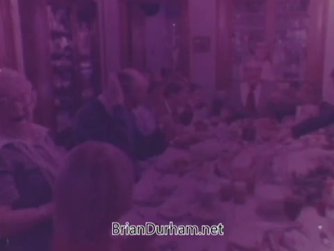 The still from the Rainbo Bread commercial features a group of people seated around a dining table, likely enjoying a meal together. The scene has a nostalgic, warm atmosphere characteristic of the 1970s. The colors are muted, contributing to the vintage feel. Participants appear to be engaged in conversation, emphasizing community and togetherness, central themes in the advertisement. The overall presentation reflects the era's style and marketing approach, focusing on family-oriented values.