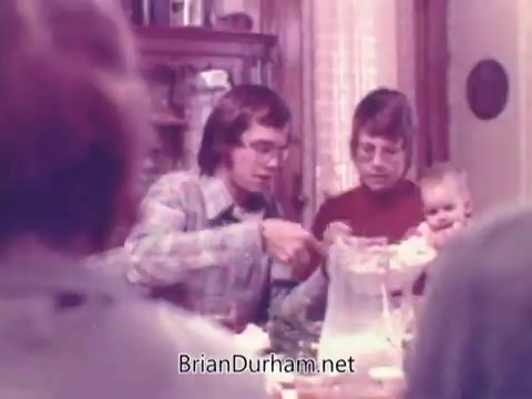 The still from the 'Rainbo Bread' commercial features a family setting typical of the 1970s. A young man in glasses and a plaid shirt serves food at a table, while a woman beside him holds a baby. The warm lighting and homey decor evoke a sense of togetherness and nostalgia related to family meals. The focus on bread suggests a connection to wholesome family dining.