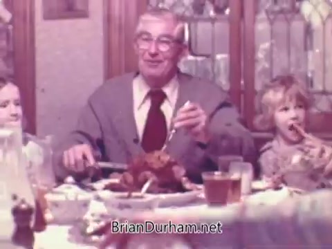 The still from the 1970s 'Rainbo Bread' commercial showcases a warm family scene at the dinner table. An elderly man, dressed in a cardigan and glasses, is serving food while sitting between two children, one of whom is eating confidently. The table is set with various dishes, creating a cozy and inviting atmosphere that highlights family togetherness. The background features wooden cabinets and soft lighting, enhancing the nostalgic mood.