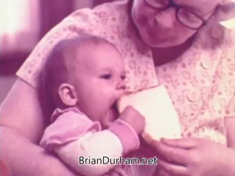 The still features a gentle scene from a 1970s Rainbo Bread commercial, showcasing an elderly woman lovingly feeding a piece of bread to a small child. The image has a nostalgic, warm color palette, emphasizing a family-oriented atmosphere. The focus is on the connection between the two, highlighting the joy of sharing and the wholesome nature of the product.