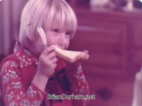 The still features a young child with blonde hair, smiling joyfully while holding a slice of bread in a playful manner, resembling a telephone. The setting appears cozy, likely in a home kitchen, reflecting a cheerful and fun atmosphere. The image is warm-toned, typical of commercials from the 1970s, emphasizing the appeal of family moments and the enjoyment of bread.