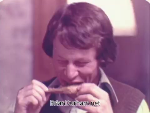 The still from the 'Rainbo Bread' commercial features a man with shaggy hair enjoying food, likely a chicken drumstick. He appears to be savoring the flavor, smiling, and engaged in the moment. The warm tones and casual setting convey a homey, inviting atmosphere typical of 1970s commercials. The focus is on the enjoyment of a meal, likely emphasizing the role of Rainbo Bread in that experience.
