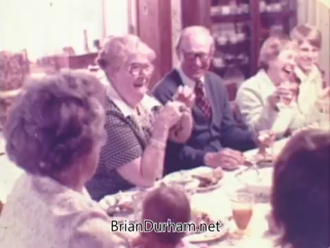 The still from the Rainbo Bread commercial features a lively family gathering around a dinner table. Several adults and a child are smiling and laughing, suggesting a warm, joyful atmosphere. The table is set with various dishes, highlighting a communal meal. This scene captures the essence of togetherness and nostalgia associated with family dining in the 1970s.