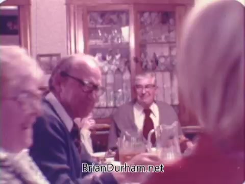 The still depicts a gathering around a dining table, featuring several older adults engaged in conversation. One man in glasses is prominently seated, with a woman beside him. The background includes a wooden hutch showcasing glassware, creating a warm, homey atmosphere typical of family gatherings in the 1970s. The overall tone suggests a sense of nostalgia and community, aligning with the themes of comfort and tradition often associated with bread advertisements from that era.