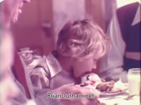 The still from the 'Rainbo Bread' commercial shows a young boy eagerly eating at a dining table with multiple people around him. The warm, soft-focus lighting suggests a cozy, family-oriented atmosphere, typical of 1970s advertisements that emphasize togetherness and the enjoyment of meals. The scene conveys a sense of nostalgia and simplicity associated with family dining.