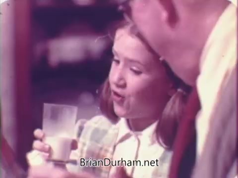 The still features a young girl holding a glass of milk while engaging with an adult, likely a parent or guardian. She appears cheerful and animated, suggesting a wholesome family moment. The setting seems domestic, typical of 1970s advertisements that emphasize family values and product enjoyment, particularly focused on promoting Rainbo Bread as a part of mealtime. The colors and fashion reflect the era's style.