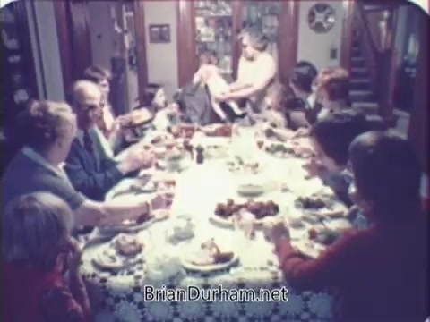 The still from the 'Rainbo Bread' commercial depicts a large, family-style dinner table filled with various dishes. A diverse group of people, including both adults and children, are gathered around the table, enjoying the meal together. The setting gives off a warm, inviting atmosphere characteristic of home-cooked meals in the 1970s. The focus on togetherness and sharing aligns with the commercial's message promoting the bread as a staple for family gatherings.