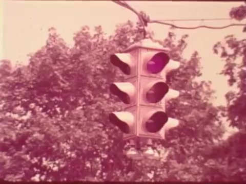 The still shows a vintage traffic light suspended above, featuring distinct signal lenses amid a backdrop of leafy trees. The colors suggest a retro aesthetic, typical of 16mm film from the era, emphasizing road safety in the context of the Aetna Public Safety Message that likely addresses pedestrian awareness.