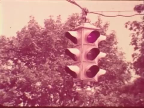 The still features a vintage traffic light with multiple signals visible against a backdrop of trees. The traffic light displays a purple light, while the other signals are either off or obscured. The overall color tone appears faded, characteristic of 16mm film. This scene likely emphasizes traffic safety in the context of Aetna's public safety message.