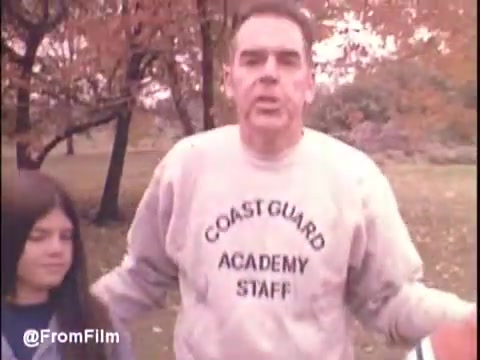 The still from the 'Otto Graham Colorectal Cancer PSA' features a man wearing a sweatshirt that reads 'COAST GUARD ACADEMY STAFF,' alongside a young girl. The setting appears outdoor, surrounded by trees showcasing autumn foliage. The overall tone suggests a personal and educational message about colorectal cancer awareness. The image has a warm, slightly faded look typical of 16mm film.