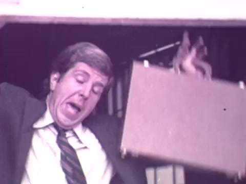The still features a man in a suit, appearing startled or distressed as he exits a building with a briefcase. His exaggerated expression suggests a comical or surprising situation. The color and quality reflect a vintage 16mm film style typical of the early 1970s, capturing the playful tone often found in advertisements of that era.
