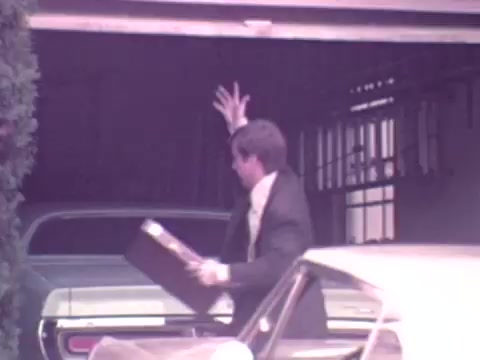 The still from the 1973 Nationwide Insurance commercial features a man in formal attire, who appears to be exiting a garage. He is holding a briefcase and raising his other hand in a gesture, possibly as a greeting or signal. In the background, there are two cars, one of which appears to be parked inside the garage. The scene has a nostalgic quality typical of the early 1970s, characterized by its color palette and clothing style.