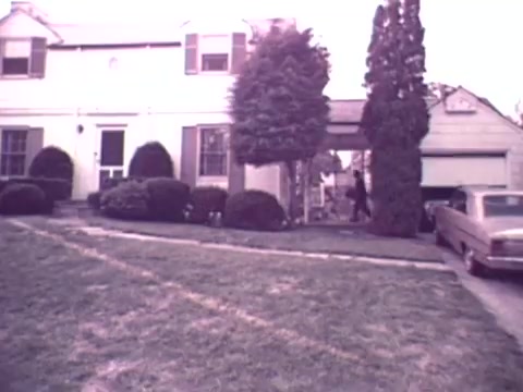 The still from the 1973 Nationwide Insurance commercial features a suburban home with a porch, neatly trimmed bushes, and a driveway leading to a garage. In the foreground, a person is seen walking towards the house. A vintage car is parked nearby, typical of the era. The overall tone is nostalgic, reflecting suburban life during the early 1970s.