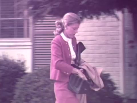 The still from the 1973 Nationwide Insurance commercial features a woman walking outside, dressed in a stylish pink suit with a white blouse. She is carrying a handbag and a light-colored coat, suggesting a professional or formal setting. The background includes greenery and a building, providing a suburban atmosphere typical of the era. The overall color tone is soft, indicative of the period's aesthetic in advertising.