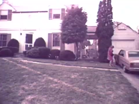 The still from the 1973 Nationwide Insurance commercial features a suburban home with a neatly manicured lawn. In the foreground, a woman in a red dress approaches a vintage car parked near the house. The scene conveys a sense of domestic life and stability, common themes in insurance advertising of that era. The overall tone is warm and inviting, typical of the period's visual style.