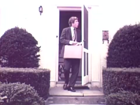 The still from the 1973 Nationwide Insurance commercial features a man dressed in a suit, stepping out of a house while carrying a briefcase. The house has a white entrance door and is flanked by neatly trimmed bushes. The overall tone of the image suggests a professional, suburban setting, typical for promoting insurance services during that era.