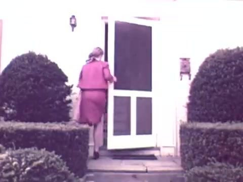 The still shows a woman in a pink outfit entering a house through a screen door. The entrance is framed by neatly trimmed hedges, suggesting a well-maintained home environment. The overall setting appears inviting, reflecting the domestic theme typical of insurance commercials from that era.