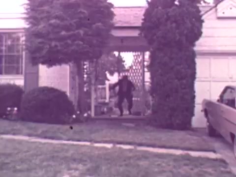 The still captures a moment from a 1973 Nationwide Insurance commercial, featuring a man emerging from a house, possibly in an exaggerated or comedic manner. The setting includes a suburban home with neatly trimmed hedges and a visible driveway. The overall tone is nostalgic, reflecting the aesthetics of that era in visual quality and color.