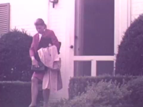 The still from the 1973 Nationwide Insurance commercial shows a woman dressed in a red outfit exiting a house. She appears to be carrying a handbag and a light-colored coat. The scene is set in a residential area, characterized by neatly trimmed hedges and a white door, conveying a suburban, domestic atmosphere. The overall tone is nostalgic and reflects the style of the early 1970s.