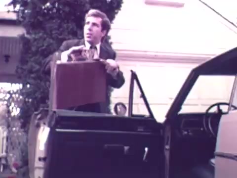 The still depicts a man in a suit standing beside an open car door, holding a briefcase and appearing concerned. He seems to be getting out of a vehicle, set in a suburban environment, with a house and greenery in the background. The scene conveys a sense of urgency or importance, typical of a narrative in commercials from that era.