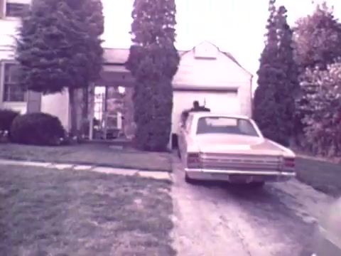 The still from the 1973 Nationwide Insurance commercial features a suburban home with a faded color palette. A vintage car is parked in the driveway, which leads up to a modest garage. The setting suggests a typical family environment of that era, emphasizing themes of security and stability. Tall trees flank the driveway, contributing to the residential ambiance.