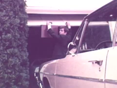The still from the 1973 Nationwide Insurance commercial depicts a man interacting with a garage door, possibly struggling to fix or open it. A vintage car is parked nearby, emphasizing the domestic setting. The scene captures a moment of everyday life, likely illustrating the importance of insurance in protecting one's home and belongings. The overall tone reflects the era's aesthetic, with a focus on relatable scenarios.