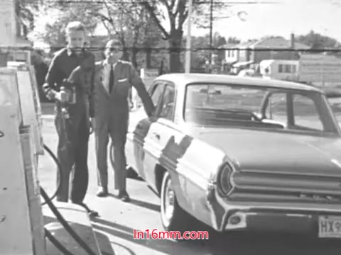 The still from 'Check for Tire Wear!' depicts two men at a gas station in the 1960s. One man, dressed casually, holds a tool, likely related to vehicle maintenance. The other, wearing a suit, stands beside a vintage car, which is parked at the pump. The scene reflects a mid-century setting, emphasizing automotive upkeep and safety.