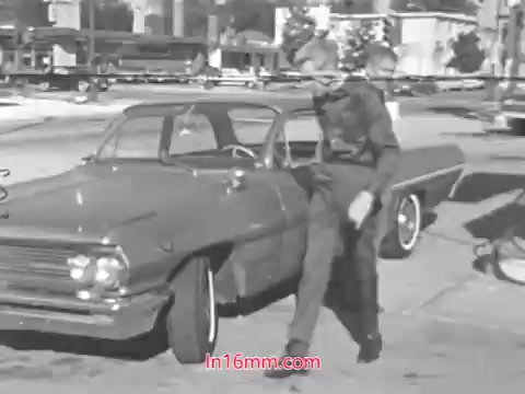 The still features a man walking away from a vintage car, likely from the 1960s, parked on a street. He appears engaged in a safety-related task, consistent with the theme of tire wear awareness. The scene conveys a sense of everyday life during that era, emphasizing vehicle maintenance and road safety. The surrounding environment includes buildings and other vehicles, indicative of a typical urban setting in that time period.