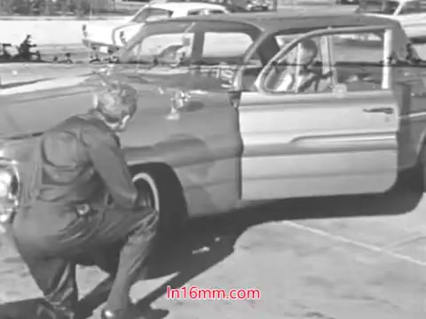The still from 'Check for Tire Wear!' features a mechanic or safety inspector examining the tire of a vintage car while a driver sits inside. The scene emphasizes vehicle safety, likely as part of a public service announcement from the 1960s. The stylistic elements, such as the black-and-white imagery and period attire, highlight the era's focus on automotive care and safety awareness.
