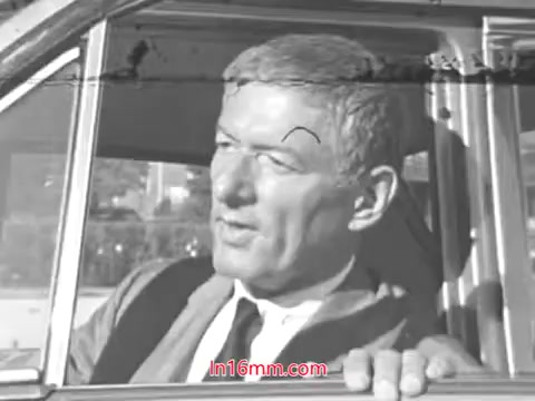 The still features a middle-aged man with light-colored hair sitting in a car, looking out of the driver's side window. He appears to be discussing or considering something, likely related to vehicle safety. The image is in black and white and has a vintage 1960s aesthetic, which aligns with the PSA's focus on awareness and caution regarding tire maintenance. The film aims to promote safety messages, indicated by the mention of the green cross symbol for safety.