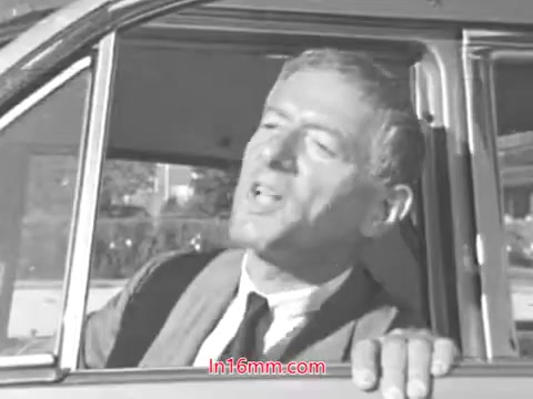 The still features a man leaning out of a car window, speaking animatedly. He appears to be conveying an important message about safety, likely related to checking tire wear, as suggested by the title 'Check for Tire Wear!' from the 1960s. His expression and posture indicate a sense of urgency and authority, emphasizing the significance of vehicle safety. The black-and-white format adds a nostalgic feel typical of public service announcements from that era.