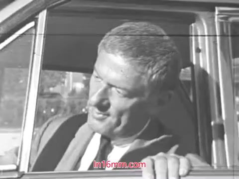 The still features a man sitting in a car, appearing to engage with someone outside the vehicle. He has a thoughtful expression and is dressed in a suit, typical of the 1960s professional attire. The setting suggests a focus on automobile safety, likely related to tire wear, as indicated by the context of the film. The black-and-white format emphasizes the period's aesthetic.