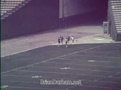 The still depicts a group of three individuals running together on a football field, surrounded by empty stadium seating. The scene conveys a focus on physical fitness, likely promoting an active lifestyle as part of the President's Council on Physical Fitness campaign from 1967. The atmosphere suggests a sense of teamwork and effort in achieving fitness goals.
