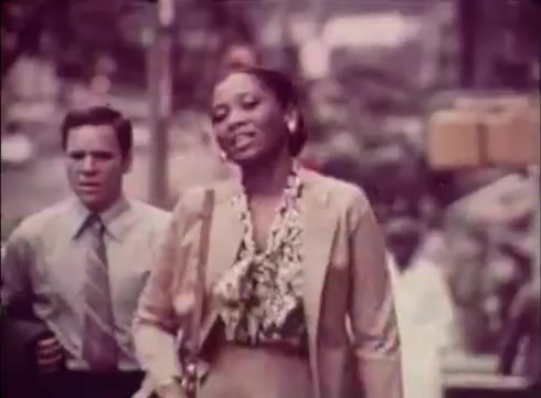The still from the 16mm print of 'Job Service Nebraska - Book Store' features a woman in a fashionable outfit, likely representing professionalism and confidence, walking with a man in a suit. The backdrop indicates a city environment, emphasizing a theme of urban life and opportunities. The color palette is muted, consistent with the vintage aesthetic of the era. This scene suggests a narrative focused on job search and career advancement, common in related commercials.