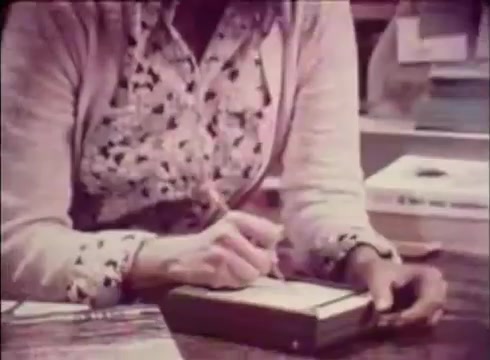 The still depicts a close-up of a woman’s hands as she writes in a notebook, likely in a bookstore setting. The focus is on the act of writing, highlighting a personal and attentive approach. The color palette suggests a warm, inviting atmosphere typical of mid-20th century commercial aesthetics. This frame captures the essence of the 'Job Service Nebraska - Book Store' theme, emphasizing service and connection in a community context.