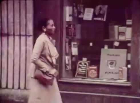 The still from 'Job Service Nebraska - Book Store' features a woman walking past a bookstore window filled with various books and promotional materials. She is dressed in a light-colored coat and appears to be smiling, suggesting a positive experience or interaction. The scene likely conveys a sense of community engagement and the importance of job services in relation to local businesses. The vintage quality of the 16mm print adds to the nostalgic tone of the advertisement.