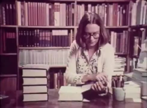 The still from 'Job Service Nebraska - Book Store' features a woman in glasses sitting at a table surrounded by stacks of books. She appears to be engaged in reading or handling a book. The backdrop consists of a well-organized bookshelf filled with various titles, suggesting a scholarly or professional environment. The scene conveys a sense of quiet contemplation and dedication to learning or career development, tying into the theme of job services and education.