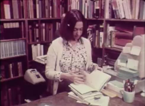 The still depicts a woman in a bookstore, standing at a counter surrounded by shelves filled with books. She appears engaged in reviewing a book while several others are stacked on the counter. The setting conveys a cozy, intellectual atmosphere typical of a community bookstore.