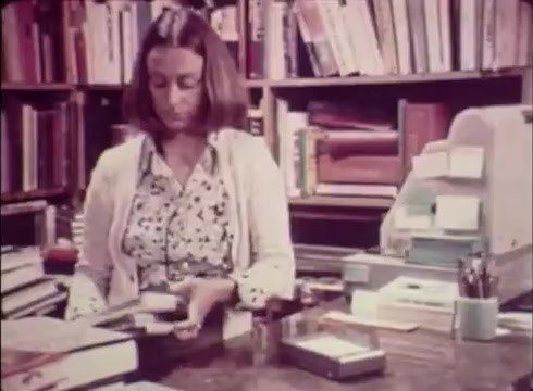 The still from 'Job Service Nebraska - Book Store' features a woman with shoulder-length hair, seated at a desk cluttered with books and office supplies. In the background, shelves filled with books are visible. The scene captures a moment of her engaged in a task, likely related to managing or organizing the materials. The overall tone suggests a focus on local employment services tied to the book store setting.
